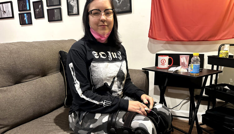 June Voros is sitting on a couch in her home. Her hands are placed on her knees and she looks towards the camera.